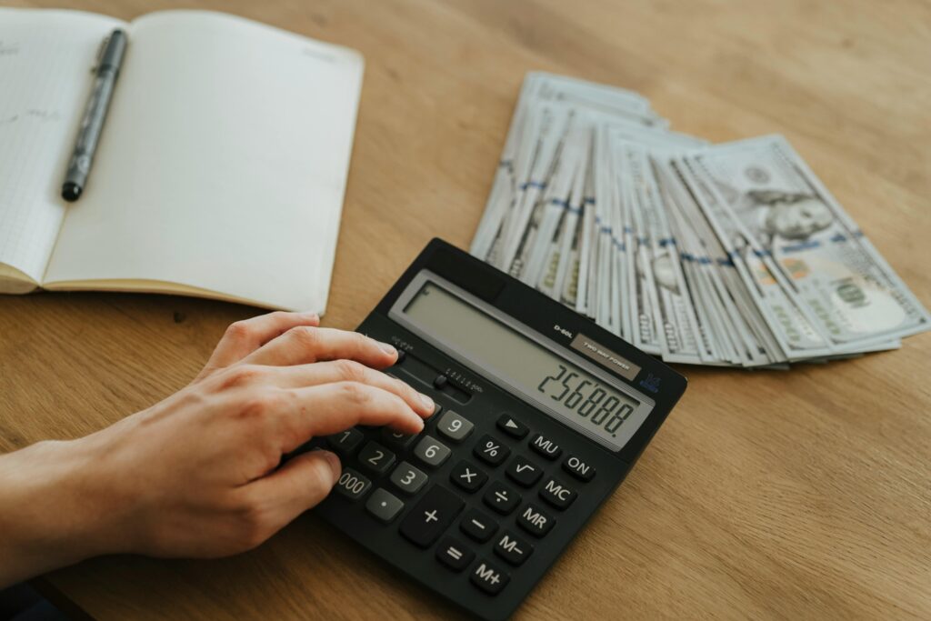 A hand calculates financial figures using a calculator with stacks of cash nearby on a wooden table.