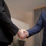 Two businessmen in suits shaking hands, emphasizing trust and partnership indoors.