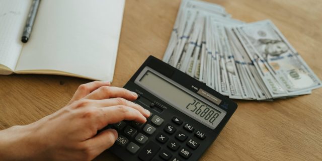 A hand calculates financial figures using a calculator with stacks of cash nearby on a wooden table.